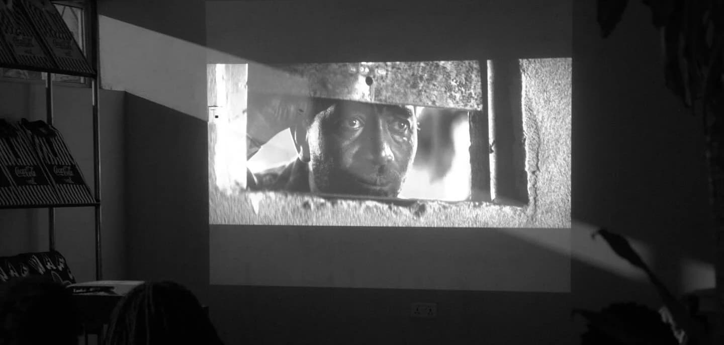 A film projected in a darkened room, a face framed in a window on screen.