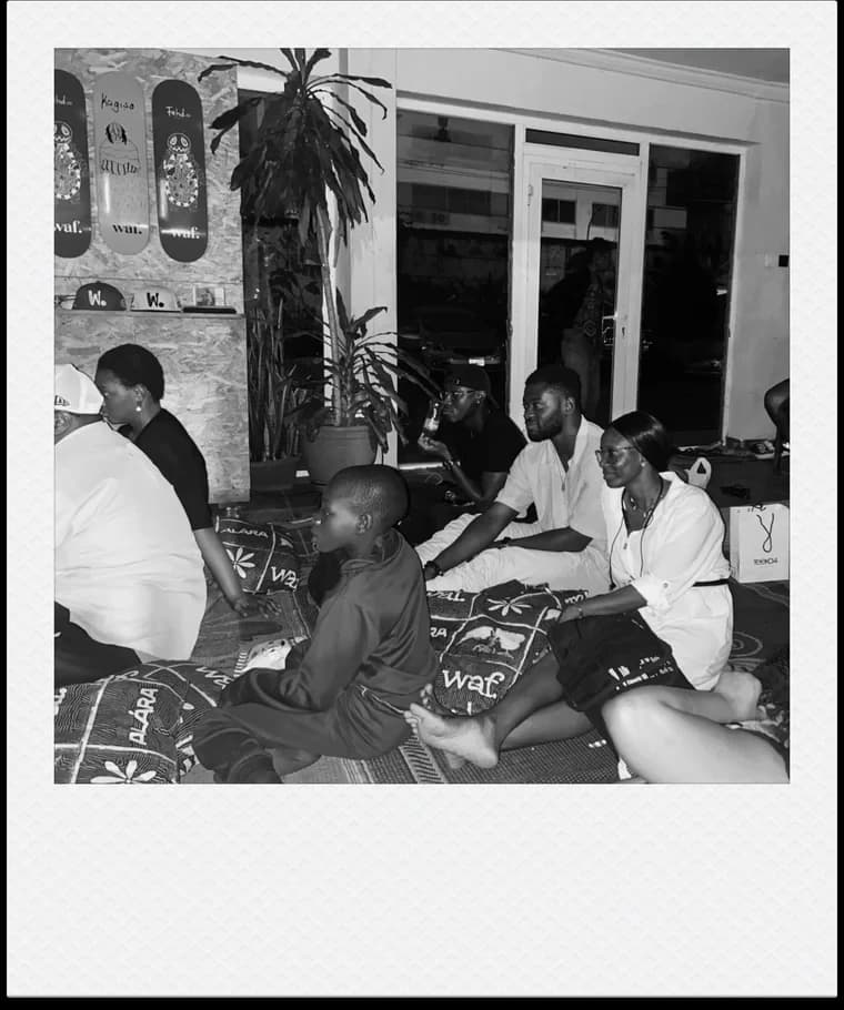 Viewers seated on patterned mats and cushions beside a tall plant and glass doors.